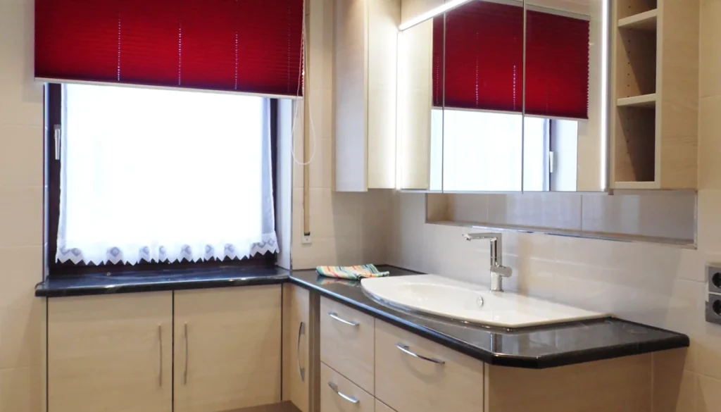 Maßgefertigte Badmöbel in Holzdekor mit schwarzer Natursteinplatte als Fensterbank und Waschtisch, rotes Plissee am Fenster.