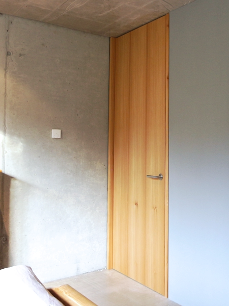 Zimmertür aus geöltem Lärchenholz in einem modernen Schlafzimmer mit Sichtbetonwand und minimalistischer Einrichtung.