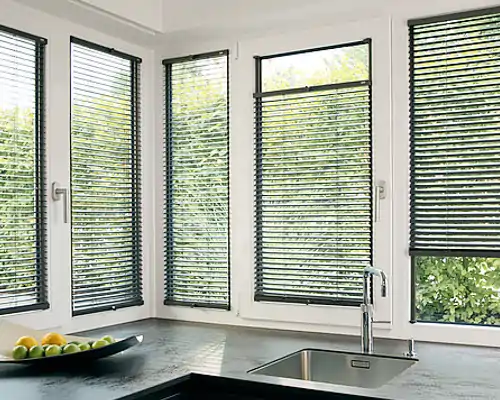 Ein Küchenschreibtisch mit MHZ Jalousien in schwarz. Die Jalousien decken die Fenster ab und bieten Sonnenschutz, während der Blick auf den grünen Garten draußen zu sehen ist