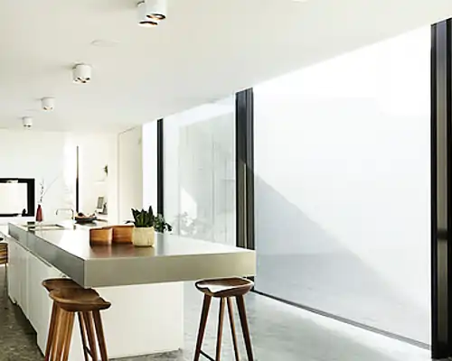 Moderne Küche mit großen Fenstern und indoor Sonnenschutz. Der Raum ist minimalistisch mit einer eleganten Kücheninsel und Hockern, Pflanzen und natürlichem Licht, das durchdringt