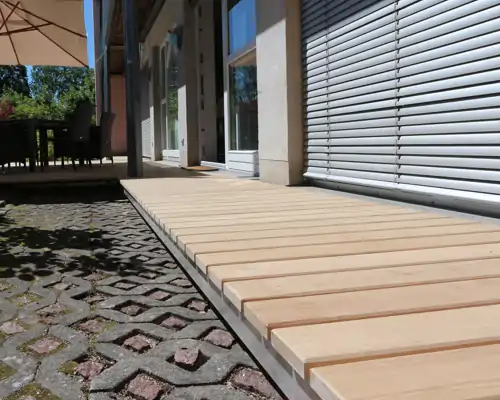 Moderne Holzterrasse mit gleichmäßig verlegten Dielen, gepflastertem Boden und sonnigem Ambiente neben einem Gebäude.