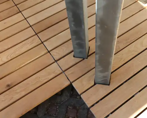 Detailansicht einer Holzterrasse mit Edelstahlstützen und gleichmäßig verlegten Dielen, die auf gepflastertem Boden ruhen.