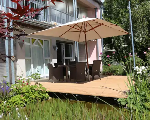 Holzterrasse mit Gartentisch, Stühlen und Sonnenschirm am Haus, direkt über einem Gartenteich mit üppiger Bepflanzung