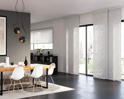 Esszimmer mit einem hellen Holztisch und weißen Stühlen mit Holzbeinen. Über dem Tisch hängen zwei schwarze Pendelleuchten mit runden Lampenschirmen. An der Wand links ist ein grauer Farbton, davor steht ein Sideboard mit Dekorationen. Rechts sind große Fenster mit weißen MHZ Flächenvorhängen, die ein dezentes, kreisförmiges Muster zeigen. Der Boden ist dunkel und glatt, ein heller Teppich liegt unter dem Tisch.