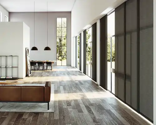 Heller, moderner Wohnraum mit großflächigen Fenstern, die mit MHZ Flächenvorhängen in dunklem Grau bedeckt sind. Rechts im Bild verlaufen die Flächenvorhänge über die gesamte Fensterfront. Links steht ein braunes Ledersofa auf einem hellen Teppich, dahinter ein Esstisch mit schwarzen Stühlen und zwei schwarzen Hängeleuchten. Der Boden besteht aus hell-dunkel gemasertem Holz.