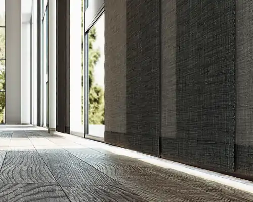 MHZ Flächenvorhang aus dunkelgrahem, leicht strukturiertem Stoff, der in mehreren Bahnen von der Decke bis zum Boden hängt. Der Vorhang steht vor großen, bodentiefen Fenstern, durch die Tageslicht einfällt. Der Boden besteht aus hellbraunem Holz mit sichtbarer Maserung.