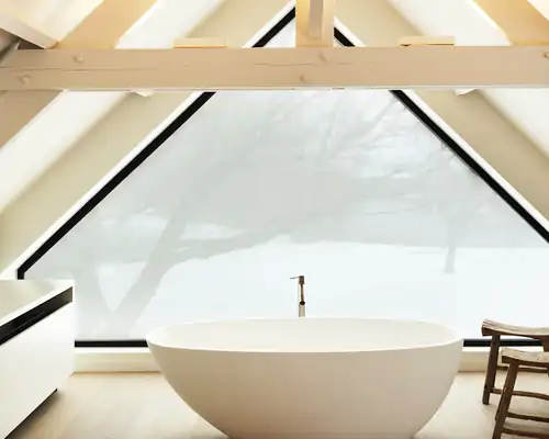 Freistehende, weiße Badewanne vor einem großen, dreieckigen Fenster mit MHZ Hafttextil SQUID Sichtschutzfolie. Im Vordergrund links ein weißer Waschtisch, rechts ein Holzstuhl. Sichtbare Holzbalken an der Decke, heller Holzboden.