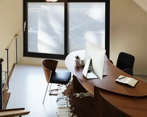 Halbrunder, dunkelbrauner Schreibtisch mit Computerbildschirm, Stiftbehältern und aufgeklapptem Notizbuch. Zwei Stühle, einer mit schwarzer Sitzfläche und Holzlehne, der andere komplett schwarz. Helle Fenster mit halbtransparenten, weißen MHZ Hafttextil SQUID Sichtschutzfolien. Heller Boden mit braun-weißer Fellmatte. Treppengeländer links aus Holz und Metall.