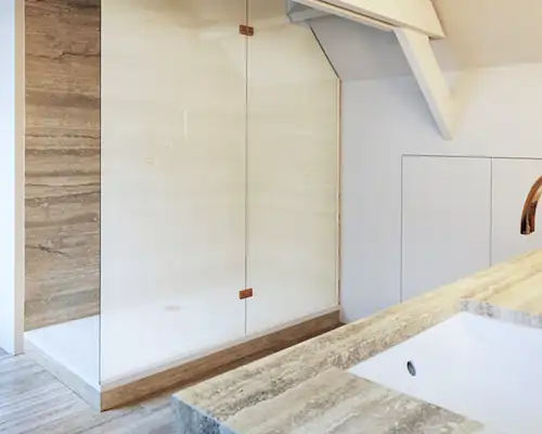 Badezimmer mit einer Glasdusche, die durch zwei kupferfarbene Beschläge an der Wand befestigt ist. Die Dusche steht auf einem Holzsockel, der zum Bodenbelag passt. Im Vordergrund ist eine Arbeitsplatte aus hellem Naturstein mit eingelassenem Waschbecken zu sehen. Die Wände sind weiß, und eine schräge Decke verläuft über der Dusche.