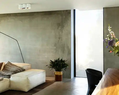 Helles Wohnzimmer mit grauer Wand und großem Fenster, das mit einem halbtransparenten MHZ Hafttextil SQUID Sichtschutz bedeckt ist. Links steht ein weißes Ledersofa mit braunem Kissen, daneben ein Topf mit grüner Pflanze. Rechts ist ein Holztisch mit schwarzem Stuhl zu sehen.