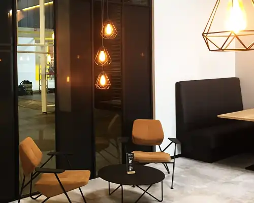 Dunkle, rahmenlose Glaswand mit drei hängenden, warm leuchtenden Pendelleuchten in geometrischer Form. Vor der Glaswand zwei beige Sessel mit schwarzen Metallgestellen und ein runder, schwarzer Tisch mit dünnen Beinen. Auf dem Tisch steht ein kleines, schwarzes Schild mit weißer Aufschrift. Rechts daneben eine schwarze Sitzbank an der weißen Wand, darüber eine große, geometrische Deckenleuchte aus schwarzem Metall.