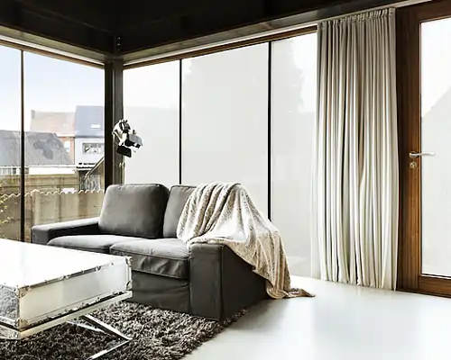 Wohnzimmer mit dunklem Sofa, auf dem eine helle Decke liegt. Große Fenster mit MHZ Hafttextil SQUID Sichtschutzfolie und hellen Vorhängen. Heller Boden, niedriger Couchtisch mit Metallgestell und dunklem Teppich. Holzrahmen um Fenster und Tür.