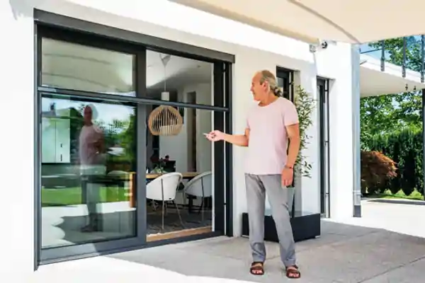 Ein Mann steht vor einer großen, bodentiefen Schiebetür mit schwarzem Rahmen, die zu einem modernen Wohnraum führt. Die Tür ist mit einem elektrischen Insektenschutzrollo ausgestattet, das halb geöffnet ist. Der Mann trägt ein helles T-Shirt, graue Hose und Sandalen und hält eine Fernbedienung in der Hand. Im Innenraum sind ein runder Esstisch mit weißen Stühlen und eine hängende Lampe aus Holz sichtbar. Im Hintergrund ist ein Garten mit Bäumen und Sträuchern zu sehen.