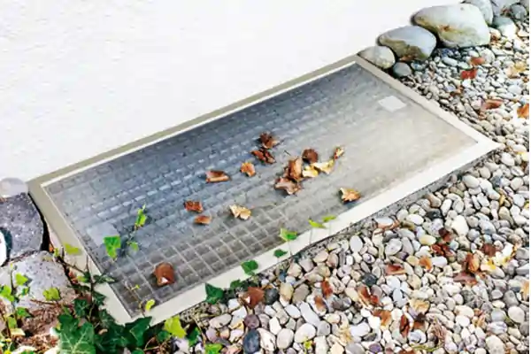 Eine rechteckige Lichtschachtabdeckung aus Metall mit feinem Gittermuster liegt auf einem Kiesbett. Auf der Abdeckung liegen verstreut trockene Blätter. Um die Abdeckung herum sind kleine Steine und etwas grüne Pflanze zu sehen.