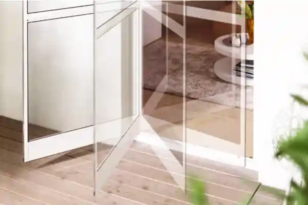 Pendeltür mit Insektenschutzgitter aus transparentem Material, montiert in einem weißen Türrahmen. Die Tür steht offen und zeigt den Übergang von einem hellen Holzboden im Außenbereich zu einem Innenraum mit Teppich und weißem Möbelstück. Im Vordergrund unscharfe grüne Blätter.
