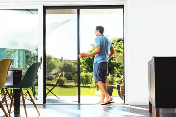 Ein Mann in blauem Poloshirt, dunklen Shorts und Flip-Flops steht in einem hellen Raum vor einer großen Glasschiebetür. Er hält den Griff eines schwarzen Plissee-Insektenschutzgitters, das halb geöffnet ist. Draußen ist ein grüner Garten mit Bäumen und einem Stuhl zu sehen. Im Raum sind ein Holztisch mit schwarzen Stühlen und ein dunkler Schrank sichtbar.