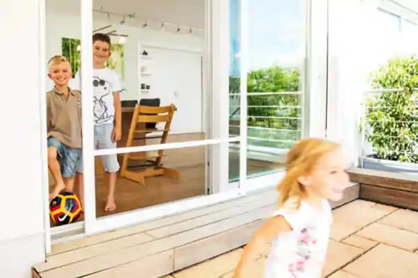 Zwei Kinder stehen hinter einer großen Schiebetür mit integriertem Insektenschutzgitter. Die Tür besteht aus Glas und hat einen weißen Rahmen. Vor der Tür läuft ein weiteres Kind auf einer Holzterrasse. Im Hintergrund sind ein Esstisch mit Stühlen und ein Balkon mit Geländer zu sehen. Die Umgebung wirkt hell und freundlich.