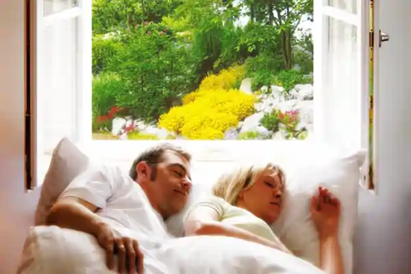 Zwei Personen liegen nebeneinander auf einem weißen Sofa vor einem geöffneten Fenster. Das Fensterrahmen ist weiß und gibt den Blick auf einen Garten mit grünen Pflanzen und gelben Blumen frei. Die Personen tragen helle Kleidung und entspannen sich in einer hellen, freundlichen Umgebung.