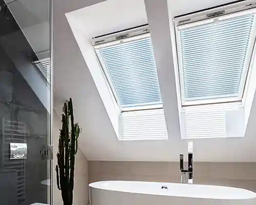 Badezimmer mit zwei Dachfenstern, die mit MHZ Jalousien ausgestattet sind. Unter den Fenstern steht eine freistehende weiße Badewanne. Links neben der Badewanne steht eine hohe, schlanke Pflanze. Rechts von der Badewanne ist ein moderner Wasserhahn installiert. Die Wände sind hell und schlicht gestaltet.