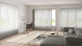 Helles Wohnzimmer mit großen Fenstern, die mit weißen MHZ Lamellenvorhängen ausgestattet sind. Die Lamellen erzeugen Schattenmuster auf dem hellen Boden. Im Raum steht ein graues Ecksofa mit Kissen, ein niedriger Holztisch mit Dekoration und ein schwarzer Klappstuhl mit Metallgestell. Die Wände sind weiß, der Raum wirkt modern und minimalistisch.