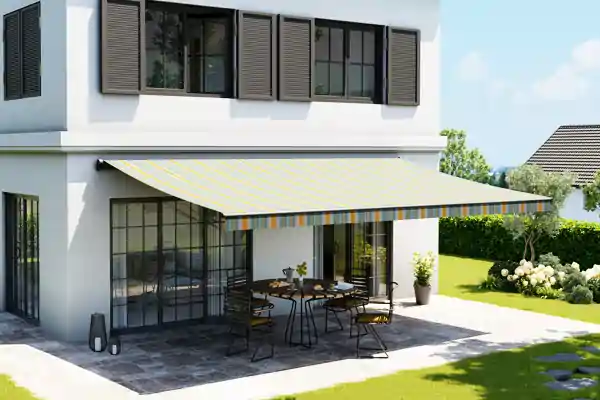 An einem weißen zweistöckigen Haus mit großen Fenstern und braunen Fensterläden ist eine Gelenkarmmarkise mit gelb-weißen Streifen von der Markisenaktion angebracht. Die Markise spendet Schatten über einer gepflasterten Terrasse, auf der ein runder Tisch mit mehreren Stühlen steht. Auf dem Tisch befinden sich eine Vase mit Blumen und weitere kleine Gegenstände. Im Hintergrund ist ein grüner Garten mit Rasen, Sträuchern und einem weiteren Haus zu sehen.