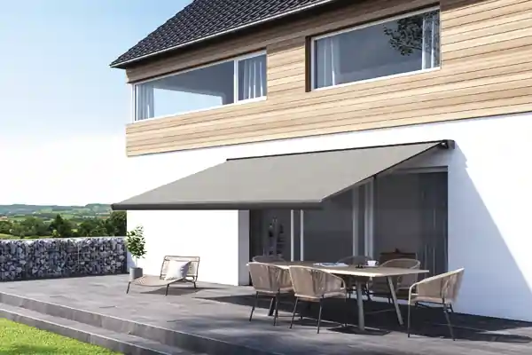 Ein modernes Haus mit einer großen Terrasse. An der Hauswand ist eine graue Kassettenmarkise von der Markisenaktion ausgefahren, die Schatten auf den Terrassenbereich wirft. Unter der Markise steht ein Esstisch mit mehreren Stühlen. Links auf der Terrasse befindet sich eine einzelne Sitzgelegenheit mit Kissen. Im Hintergrund ist eine niedrige Steinmauer und eine grüne Landschaft zu sehen.