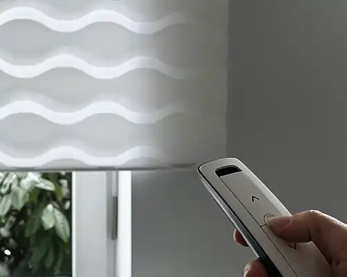 Eine Hand hält eine schlanke, weiße Somfy IO Fernbedienung mit zwei Pfeiltasten, die nach oben und unten zeigen. Im Hintergrund ist ein weißes Rollo mit wellenförmigem Muster zu sehen, das teilweise vor einem Fenster hängt. Durch das Fenster sind grüne Blätter von Pflanzen sichtbar.