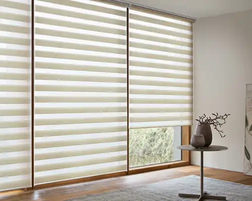 Zwei große Fenster mit MHZ Doppelrollos, die aus abwechselnd transparenten und blickdichten horizontalen Streifen bestehen. Die Rollos sind halb heruntergelassen, sodass durch die transparenten Streifen ein Blick nach draußen möglich ist. Rechts neben den Fenstern steht ein runder Beistelltisch mit einem dunkelgrauen, bauchigen Keramikvase, in der ein kahler Zweig steckt. Der Boden ist mit einem hellen Teppich bedeckt, die Wände sind weiß.