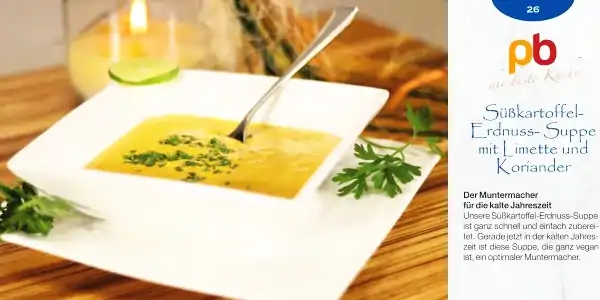 Weiße quadratische Schale mit gelber Süßkartoffel-Erdnuss-Suppe, garniert mit grünen Kräutern, daneben Limettenscheibe und frische Kräuter.