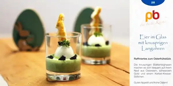 Zwei Gläser mit grünem Püree, darauf ein gekochtes Ei, garniert mit Kresse und einem knusprigen Langohren-Keks, auf einem Holztisch vor grünen Osterdeko-Eiern.