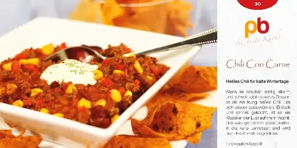 Weiße Schale mit Chili con Carne, garniert mit saurer Sahne und Mais, daneben Tortilla-Chips in einer Schale.