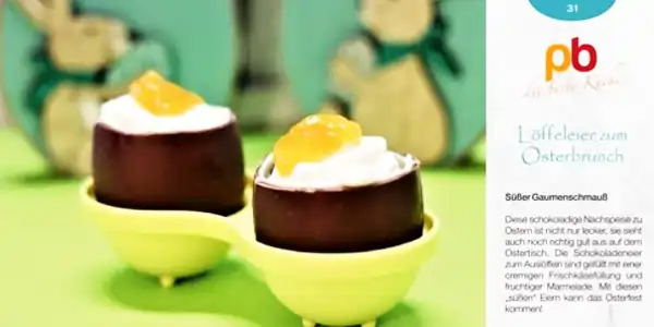 Zwei Schokoladeneier mit weißer Füllung und gelber Marmelade, serviert in gelben Löffeleiern vor grünem Hintergrund mit Osterdeko.