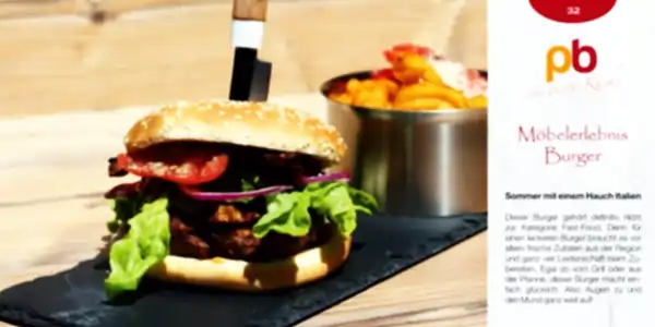 Ein Burger mit Sesambrötchen, gefüllt mit Salat, roten Zwiebelringen, Tomatenscheiben und einer dicken, gebratenen Fleischscheibe. Im Hintergrund steht ein Metallbecher mit Pommes frites. Ein Messer steckt senkrecht in den Burger. Der Untergrund ist eine helle Holzfläche. Rechts oben ist ein Teil einer Rezeptkarte mit dem Titel 'Möbeleerlebnis Burger' und dem Text 'Sommer mit einem Hauch Italien' zu sehen.