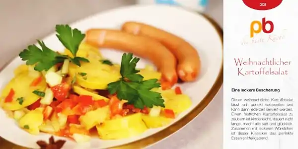 Auf einem weißen Teller mit braunem Rand liegt ein weihnachtlicher Kartoffelsalat, der aus gewürfelten Kartoffeln, roten Paprikastücken und Zwiebeln besteht. Der Salat ist mit frischer Petersilie garniert. Daneben liegen zwei helle Würstchen. Im Hintergrund ist ein Rezeptkartendesign mit dem Logo "pb die beste Küche" und dem Titel "Weihnachtlicher Kartoffelsalat" zu sehen. Darunter steht der Text "Eine leckere Bescherung" und eine kurze Beschreibung des Gerichts.