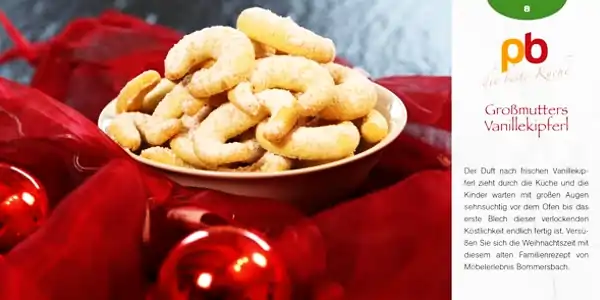 Schale mit Vanillekipferln, kleinen halbmondförmigen Keksen, auf rotem Stoff drapiert. Im Hintergrund unscharfe rote Weihnachtskugeln. Rechts daneben Text mit dem Titel 'Großmutters Vanillekipferl' und Beschreibung, die den Duft und die Vorfreude auf die Kekse beschreibt. Oben rechts ein Logo mit den Buchstaben 'pb' und dem Schriftzug 'die beste Küche'.