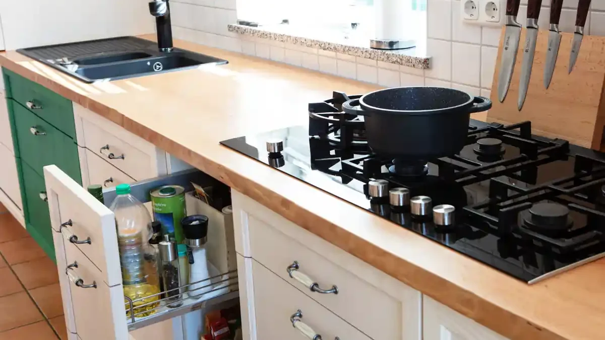 Küchenarbeitsplatte aus Buche mit sichtbarer Waldkante, darauf ein schwarzes Gaskochfeld mit fünf Brennern und einem schwarzen Topf. Links ein schwarzes Spülbecken mit Armatur. Unter der Arbeitsplatte weiße Schränke mit Metallgriffen und Esche Fronten, ein Schublade ist geöffnet und zeigt ein ausziehbares Regal mit verschiedenen Flaschen und Behältern. An der Wand rechts Messer in einem Messerblock. Die Rückwand ist mit weißen quadratischen Fliesen verkleidet.