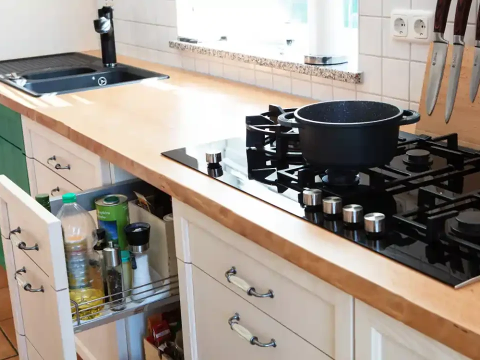Küchenarbeitsplatte aus Buche mit sichtbarer Waldkante, darauf ein schwarzes Gaskochfeld mit fünf Brennern und einem schwarzen Topf. Links ein schwarzes Spülbecken mit Armatur. Unter der Arbeitsplatte weiße Schränke mit Metallgriffen und Esche Fronten, ein Schublade ist geöffnet und zeigt ein ausziehbares Regal mit verschiedenen Flaschen und Behältern. An der Wand rechts Messer in einem Messerblock. Die Rückwand ist mit weißen quadratischen Fliesen verkleidet.