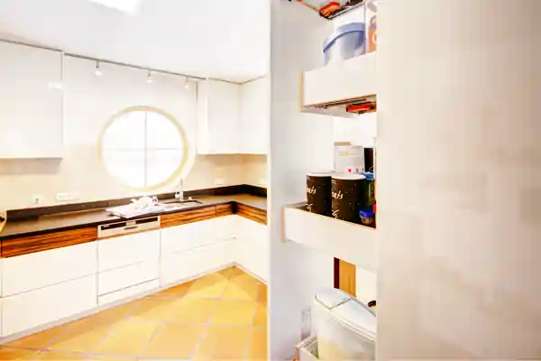 Helle Schreinerküche mit weiß lackierten Fronten und Holzarbeitsplatte. Rechts im Vordergrund ein offener Apothekerauszug mit mehreren Fächern, darin verschiedene Vorratsbehälter und Dosen. Im Hintergrund ein rundes Fenster über der Spüle, die in die Arbeitsplatte integriert ist. Die Küche ist modern und minimalistisch gestaltet, der Boden besteht aus hellen Fliesen.