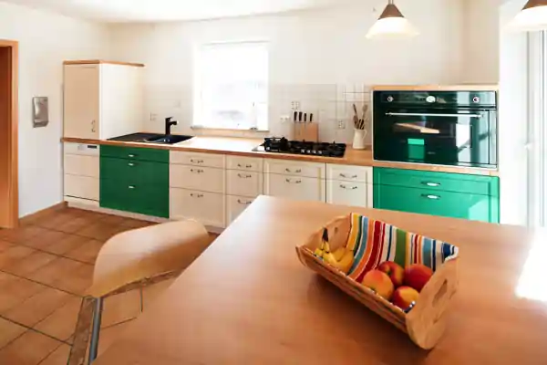 Küche mit massiven Esche-Holzfronten in grün und naturfarbenem Holzarbeitsplatten. Unter der Arbeitsplatte befinden sich grüne Schranktüren und Schubladen mit silbernen Griffen. Rechts ist ein eingebauter Backofen in schwarzer Farbe. Auf der Arbeitsplatte steht ein Messerblock und ein weißer Behälter mit Küchenutensilien. Links ist ein schwarzes Spülbecken mit Armatur. In der Mitte des Raums steht ein Holztisch mit einem Korb, der mit einem bunten Tuch ausgelegt ist und verschiedene Früchte enthält. Der Boden ist mit braunen Fliesen belegt, und an der Decke hängt eine weiße Pendelleuchte. Zwei Fenster lassen Tageslicht in den Raum.