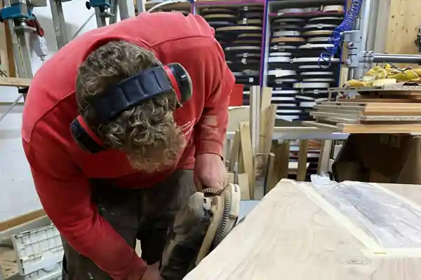 Pius Bommersbach trägt einen roten Pullover und einen schwarzen Gehörschutz. Er arbeitet in der Schreinerei in Schongau an einer hellen Rüster Holzarbeitsplatte, die auf einem Arbeitstisch liegt. Im Hintergrund sind Regale mit vielen runden Holzscheiben und weiteren Werkstattmaterialien zu sehen.