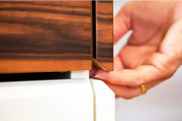 Nahaufnahme einer Schreinerküche mit Griffleiste aus dunklem Holz. Eine Hand mit goldfarbenem Ring greift an die Griffleiste, die bündig in die Front integriert ist. Unter der Holzfront ist ein weißes Element sichtbar.