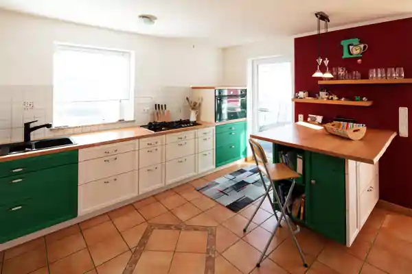 Küche mit Kochinsel, die Fronten der Unterschränke sind grün und weiß lackiert. Arbeitsplatten aus Holz, die Kochinsel hat eine Holzarbeitsplatte mit zwei Stühlen. An der Wand hinter der Kochinsel sind zwei Holzregale mit Gläsern, Tassen und Dekoration. Die Wand dahinter ist rot gestrichen. Fenster und Tür lassen Tageslicht in den Raum. Boden aus terrakottafarbenen Fliesen mit Muster in der Mitte.