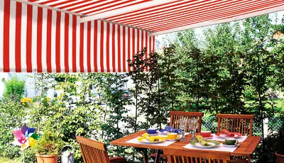 Eine Terrasse mit einer rot-weiß gestreiften Tragrohrmarkise, die Schatten spendet. Darunter steht ein Holztisch, der mit buntem Geschirr, Gläsern und einer Kaffeekanne gedeckt ist. Um den Tisch herum befinden sich Holzstühle mit Rückenlehnen. Im Hintergrund sind grüne Pflanzen und Bäume zu sehen, die eine natürliche Begrenzung bilden. Die Szene wirkt einladend und zeigt eine gemütliche Sitzgelegenheit im Freien.