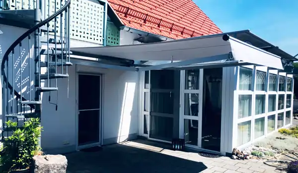An einem weißen, eingeschossigen Haus mit roten Dachziegeln ist eine weiße MHZ Tragrohrmarkise Classic Maxima ausgefahren, die den Eingangsbereich und das angrenzende Esszimmer beschattet. Die Markise ist rechteckig und wird von zwei seitlichen Armen getragen. Rechts neben der Markise befindet sich ein Wintergarten mit großen Fenstern. Links führt eine weiße Außentreppe mit Geländer nach oben. Der Boden besteht aus grauen Pflastersteinen, und im Vordergrund sind einige Pflanzen und Steine zu sehen.