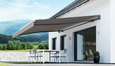 An der weißen Fassade eines modernen Hauses ist eine ausfahrbare, rechteckige Tragrohrmarkise in einem gedeckten Braunton montiert. Die Markise ist vollständig ausgefahren und spendet Schatten über einer Terrasse, auf der sich ein weißer Tisch mit vier Stühlen befindet. Die Terrasse ist mit hellen Fliesen ausgelegt, und neben der großen Glasschiebetür steht ein großer, dunkler Blumentopf mit einer grünen Pflanze. Im Hintergrund sind grüne Bäume und ein weiter Blick auf eine hügelige Landschaft unter einem hellblauen Himmel zu sehen.