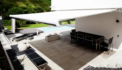 Eine weiße, ausfahrbare Kasettenmarkise vom Typ MHZ Vegas spendet Schatten über einer Terrasse. Die Terrasse ist mit großen, quadratischen Steinplatten ausgelegt und mit einem schwarzen Esstisch sowie mehreren schwarzen Stühlen ausgestattet. Im Vordergrund steht eine schwarze Liege mit silberfarbenem Gestell. Im Hintergrund ist ein Swimmingpool zu sehen, umgeben von einer gepflegten Grünfläche und Bäumen. Die Markise ist an der weißen Hauswand befestigt und erstreckt sich weit über die Terrasse hinaus.