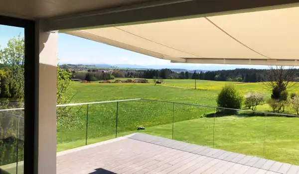 Eine hellbeige Markise spannt sich über einen Balkon mit braunen Terrassendielen aus Holz. Die Markise ist an der Decke befestigt und bietet Schatten. Im Hintergrund erstreckt sich eine grüne Wiese mit einzelnen Büschen und Bäumen, dahinter sind Hügel und ein blauer Himmel mit wenigen Wolken zu sehen.