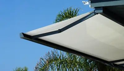 Eine hellgraue, rechteckige Kasettenmarkise mit schwarzem Rahmen, die an einer Decke montiert ist. Im Hintergrund sind grüne Palmenblätter vor blauem Himmel zu sehen.