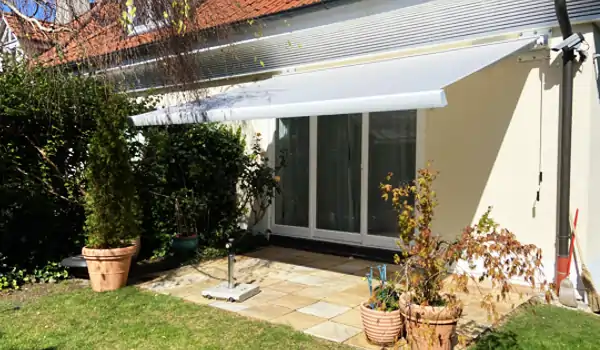 Eine hellgraue MHZ-Markise art_01 ist über einer Terrasse vor einer Küche ausgefahren und spendet Schatten. Die Terrasse besteht aus quadratischen Steinplatten, die an ein Haus mit hellen Wänden angrenzen. Vor der Terrasse stehen mehrere große Blumentöpfe mit verschiedenen Pflanzen, darunter ein hoher, grüner Strauch und weitere Pflanzen mit gelblichen Blättern. Im Hintergrund ist ein Teil des Dachs mit roten Ziegeln zu sehen. Die Markise ist an der Hauswand befestigt und erstreckt sich horizontal über die gesamte Breite der Terrasse.