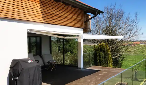 Ein modernes Wohnzimmer mit einer weißen MHZ-Markise, die über der Terrasse ausgefahren ist und Schatten spendet. Die Terrasse ist mit dunklen Bodenplatten belegt und mit einer Glasbrüstung versehen. Auf der Terrasse stehen zwei dunkelgraue Stühle und ein kleiner Tisch. Links auf der Terrasse befindet sich ein mit schwarzer Abdeckung geschützter Grill. Das Haus hat eine weiße Fassade mit einem oberen Bereich aus Holzverkleidung. Im Hintergrund sind kahle Bäume und eine grüne Wiese zu sehen.